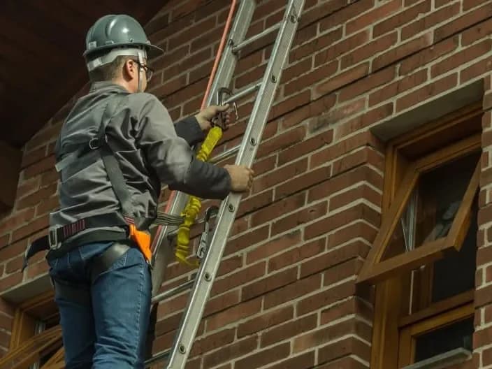 Técnico da Ecoinset, com capacete e cinto de segurança, sobe uma escada encostada na fachada de tijolos de uma casa.