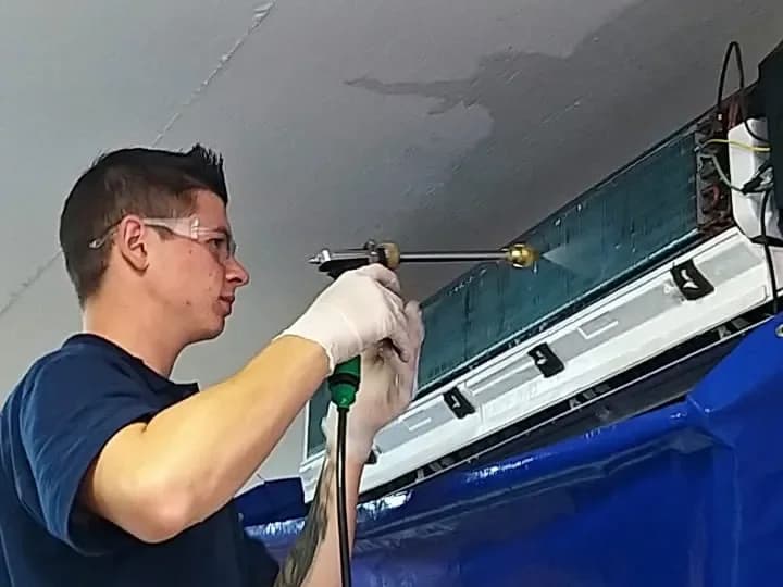 Técnico da Ecoinset, de óculos e luvas, aplica jato de limpeza na serpentina de um ar-condicionado protegido por uma capa de coleta.